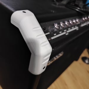 May include: A white, 3D-printed corner handle attached to a black amplifier. The handle has a curved design with three recessed finger grips and two mounting holes. The amplifier has multiple knobs and a control panel.