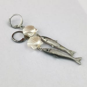 May include: Silver-toned earrings featuring a seashell and fish design. The earrings have a lever-back closure. The fish are detailed with fins and eyes. The seashells are white and have a textured surface.