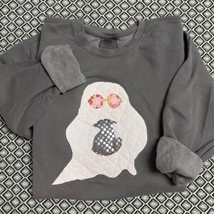 May include: A gray sweatshirt with a white ghost applique. The ghost has pink and yellow floral sunglasses and a sequined body. The sleeves are cuffed, and the sweatshirt is displayed on a patterned surface.