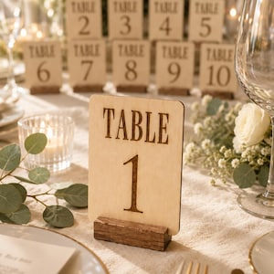 Puede incluir: Un letrero de mesa rectangular de madera clara con esquinas redondeadas, que presenta el texto "TABLE 1" grabado con láser en una fuente oscura. El letrero se mantiene en posición vertical mediante una base rectangular de madera marrón oscura.
