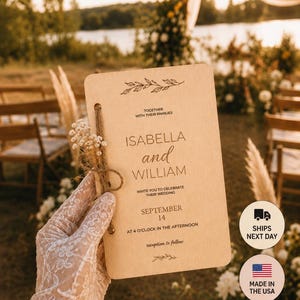 Peut inclure: Une carte d'invitation de mariage rectangulaire en bois clair aux coins arrondis, avec un texte imprimé en noir indiquant Isabella and William, September 14, et un petit bouquet de fleurs blanches séchées attaché avec de la ficelle.