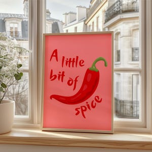 Op de afbeelding: Een roze ingelijste print met een rode chilipeper en de tekst "A little bit of spice" in rood.