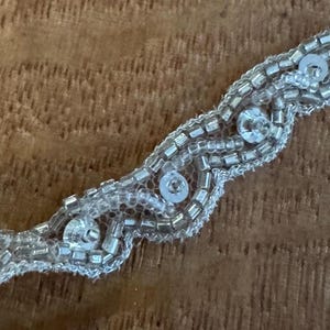 May include: A silver beaded trim with a wavy design. The trim is made of small, rectangular beads and larger, round sequins, all in a metallic silver colour. The trim is on a light brown wooden surface.