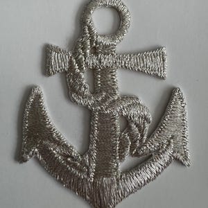 May include: A silver embroidered anchor patch with the text "Iron on Motif" at the top. The anchor is detailed with textured stitching, and the design is set against a white background. The patch is likely for nautical-themed crafts.