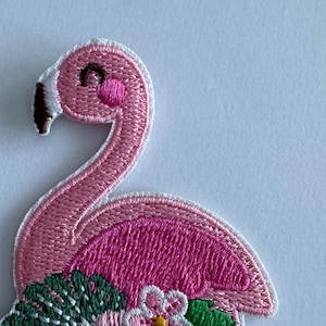 May include: Embroidered patch of a pink flamingo with a white border. The flamingo has a black beak, a rosy cheek, and a smiling expression. Green leaves and pink flowers are at the base. Ideal for customising clothing and accessories.