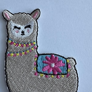 May include: Embroidered patch of a grey llama with a smiling face and a colourful beaded necklace. The llama has a blue and pink saddle blanket with a pink flower design. The patch has a black border.