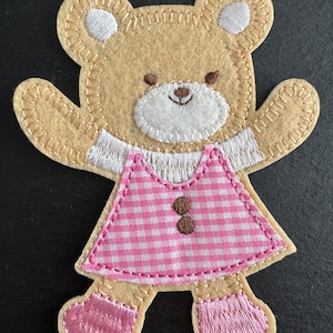 May include: A tan felt teddy bear applique with white embroidered details on the ears and chest. The bear wears a pink and white gingham dress with two brown button details and pink embroidered shoes. The applique is stitched with a contrasting thread.