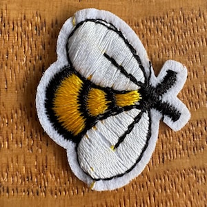 May include: Embroidered bee patch with white, yellow, and black details. The bee features white wings, a yellow and black striped body, and black antennae. The patch is outlined in black and has a white felt backing.