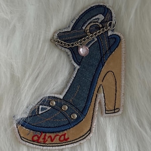 May include: Embroidered patch of a high-heeled shoe. The shoe has denim-style fabric, a tan heel, and the word "diva" in red script. A silver chain with a pink gem hangs from the ankle strap.
