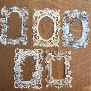 May include: Five decorative picture frames in various shapes, including rectangular and oval. The frames are white and gold, decorated with floral embellishments such as roses and daisies. They are arranged on a wooden surface.