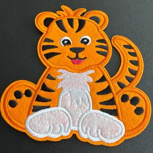 May include: An orange tiger patch with black stripes, white paws, and a pink tongue. The embroidered patch features a cartoon tiger design, ideal for decorating clothing or accessories. The tiger is sitting with its legs spread out.
