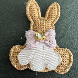 May include: A gold-coloured bunny-shaped applique with a light pink bow and a white mesh skirt. The bow features a square, light green gem. The applique is likely for clothing or craft projects.