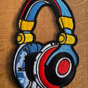 May include: Embroidered headphone patch with a black border. The design incorporates blue, red, yellow, and white stitching. The earcups have black centres with red and white details. The top arch is blue with red and black stripes.