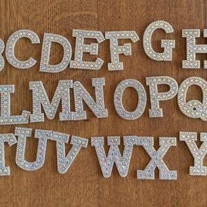 May include: A collection of silver alphabet letters, each embellished with tiny, iridescent rhinestones, rests on a wooden surface. The letters A to Z are visible, presenting a sparkling, decorative font. These letters are suitable for crafts and customisation.