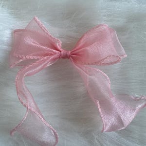 May include: A pale pink bow made from a sheer material. The bow features a traditional design with looped ends and long, flowing ribbons. The edges are finished with matching pink stitching.