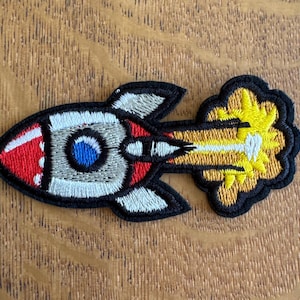 May include: Embroidered patch of a cartoon rocket ship in red, white, grey, and blue, with a yellow exhaust trail. The patch has a black border and is likely for clothing or accessories.