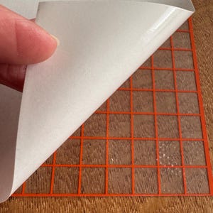 May include: A sheet of white adhesive vinyl being peeled back to reveal an orange grid pattern. The grid is on a wooden surface. The vinyl is being peeled back by a person's fingers.