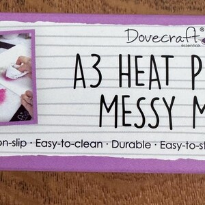 以下が含まれることがあります： DovecraftのA3 Heat Proof Messy Mat。紫色の縁取りのある白いマットで、使用中のマットの画像が特徴です。マットのテキストには「Non-stick」、「Non-slip」、「Easy-to-clean」、「Durable」、「Easy-to-store」、「Multi use」が含まれています。