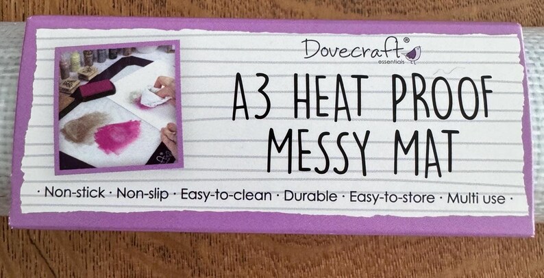 以下が含まれることがあります： DovecraftのA3 Heat Proof Messy Mat。紫色の縁取りのある白いマットで、使用中のマットの画像が特徴です。マットのテキストには「Non-stick」、「Non-slip」、「Easy-to-clean」、「Durable」、「Easy-to-store」、「Multi use」が含まれています。