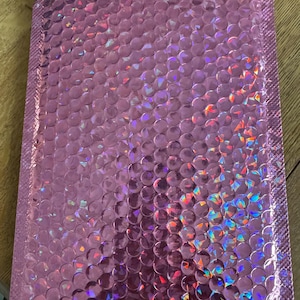 May include: A pink, iridescent bubble mailer. The mailer is rectangular with a sealed top edge. The surface is covered in small, raised, circular bubbles. The bubbles reflect a rainbow of colours. The mailer is for shipping small items.