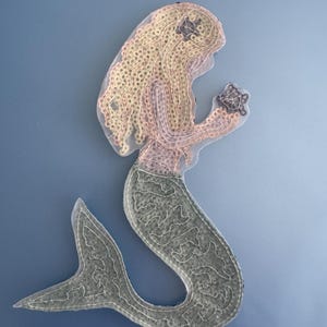May include: A fabric mermaid applique against a light blue backdrop. The mermaid features blonde hair, a pink torso, and a green tail. She holds a small, dark-coloured object. The applique is created with a stitched design, suitable for crafts.