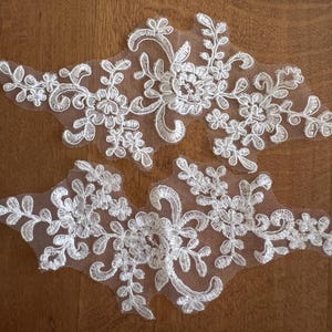 May include: Two white lace appliques featuring floral and leaf patterns. The appliques are crafted from delicate, embroidered lace, set on a sheer mesh backing. The intricate details include flowers, leaves, and swirling designs, ideal for embellishing garments or crafts.