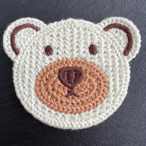 May include: A cream-coloured, knitted bear face applique with brown and tan details. The bear's head is cream with brown ear and eye accents. The snout is tan, and the nose is brown. Ideal for craft projects, this handmade applique adds a charming touch.