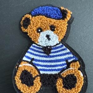 May include: Embroidered teddy bear patch with a blue sailor hat and a blue and white striped shirt. The bear has a tan body, black accents, and a white muzzle. The patch is on a black background.