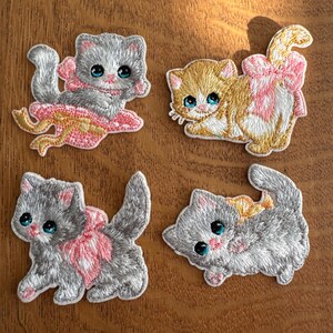 May include: Four embroidered kitten patches. Two grey kittens and one ginger kitten have pink bows, while the fourth grey kitten has a yellow bow. Each kitten has blue eyes and is in a different pose.