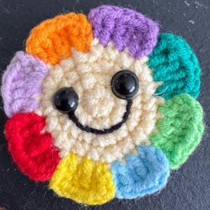 May include: A handmade crochet flower with a smiling face. The flower has a cream-coloured centre and colourful petals in shades of orange, purple, green, blue, yellow, and red. Black plastic eyes and a black stitched smile complete the design.