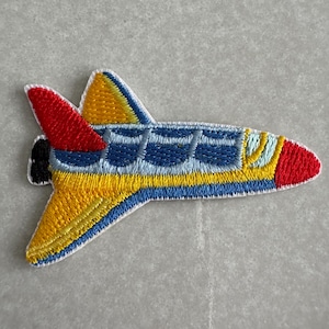 May include: Embroidered patch of a colourful space shuttle. The patch features a red nose, yellow and blue wings, and a blue body with yellow and light blue accents. The design is outlined in white.
