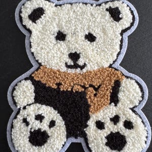 May include: A white teddy bear patch with black features and paw prints. The bear has a brown scarf and black arms and torso. The patch is outlined in light blue.