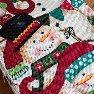 May include: Close-up of a festive fabric with a repeating pattern of snowmen. The snowmen have black top hats, red scarves, and green scarves. The background is a light beige colour, and there are small decorative elements.