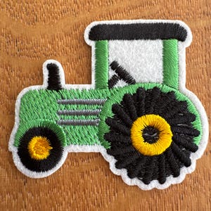 May include: Embroidered patch of a green tractor with black and yellow wheels. The tractor has a white cabin and black accents. The patch is outlined in white and is likely for clothing or accessories.