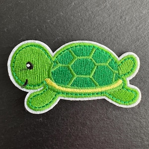 May include: A green embroidered turtle patch with a white border. The turtle has a smiling face, a yellow stripe, and a textured shell design. The patch is designed for clothing or accessories.