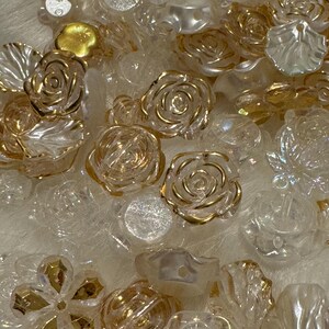 May include: A selection of clear and gold-coloured rose-shaped beads and embellishments. Some beads have gold accents, while others are translucent. The beads vary in size and shape, creating a textured and decorative assortment.