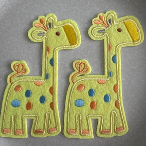 May include: Two yellow felt giraffe patches with orange, blue, and yellow accents. The giraffes have a yellow body with orange and blue spots, a yellow head with a yellow ear, and a decorative orange bow. Ideal for craft projects.