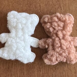 May include: Two small, handmade teddy bear ornaments. One is white, the other is a light brown colour. Both have a fluffy, textured appearance and are positioned side by side, as if holding hands. The bears are set against a wooden background.