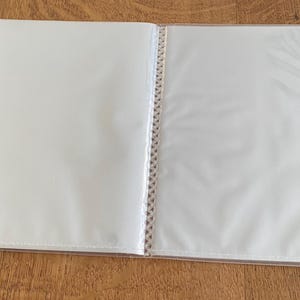 May include: An open, clear plastic photo album with multiple transparent sleeves. The album is lying on a wooden surface. The sleeves are empty and ready to hold photos or other documents.