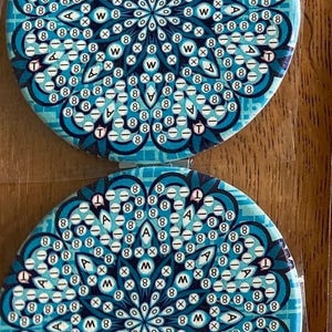 May include: Two circular coasters featuring a teal and white mandala pattern. The design incorporates the letters "A", "W", and "T", alongside the number "8". These coasters are made from a glossy material, suitable for home use.