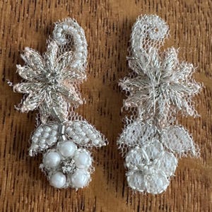 May include: Two symmetrical appliques with intricate detailing. Each features a silver-coloured, star-shaped design, delicate lace, and clusters of white beads. The appliques are set against a wooden background, highlighting their delicate craftsmanship.