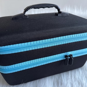 May include: A black rectangular carrying case with a black handle and two light blue zips. The case is made of a textured material and has a rectangular shape. The zips are parallel and run across the front of the case.