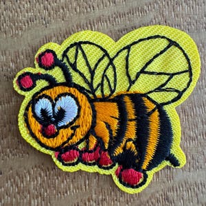 May include: Embroidered bee patch on a yellow background. The bee features a smiling face, orange body with black stripes, and red accents. The wings are black with a detailed vein pattern.
