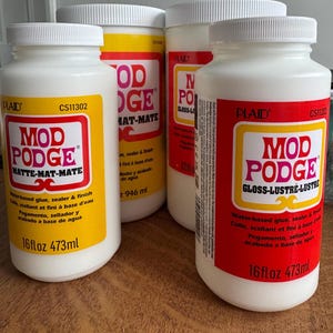 May include: Four white bottles of Mod Podge craft glue. Two bottles have yellow labels with red text, and two have red labels with yellow text. Each bottle contains 473ml of water-based glue, sealer, and finish.