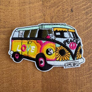 May include: Embroidered patch depicting a vibrant VW bus with a retro design. The bus is adorned with the words "LOVE" and "PEACE", a sunflower, and a peace symbol. The patch has a white border and the words "LOVE BUS" are at the bottom.