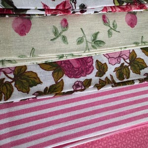 May include: Close-up of fabric samples with floral and striped designs. The top swatch displays pink roses on white. Other fabrics show pink roses on cream, pink and white stripes, and a pink floral pattern. Ideal for craft projects.