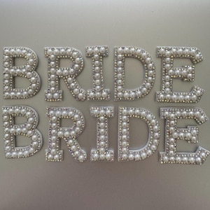 May include: Two sets of the word "BRIDE" spelled out in individual letters. Each letter is outlined with small, round, white pearls and clear rhinestones. The letters are on a light grey background.