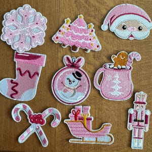 May include: A selection of pink and white Christmas-themed embroidered patches. Designs include a snowflake, Christmas tree, Santa Claus, stocking, snowman bauble, mug with gingerbread man, candy canes, sleigh, and a nutcracker.