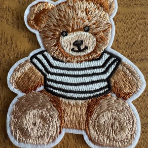 May include: Embroidered teddy bear patch with brown fur and a striped black and white shirt. The patch has a white border and is designed for application on clothing or accessories. The bear has a friendly expression.