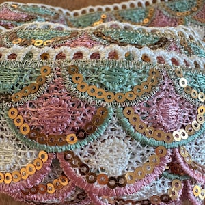 May include: Close-up of decorative trim with a scalloped edge. The trim features a combination of white, light green, and pink embroidery, accented with gold sequins. The design is intricate and detailed, suitable for embellishing clothing or craft projects.
